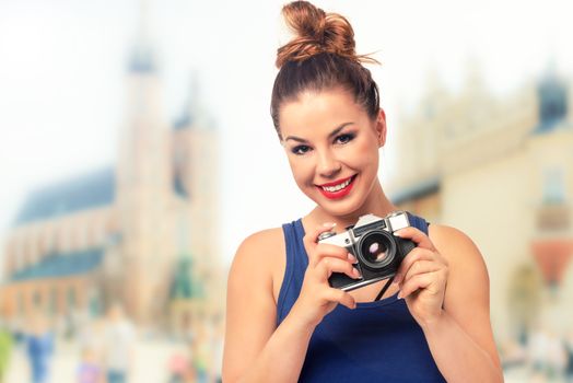 Vacation photographer concept - beautiful and attractive woman holding a retro SLR camera and smiling on blurred background of a historic city (vintage effect).