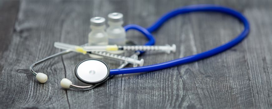 A blue medical stethoscope, syringes and vials sit on a wooden surface.