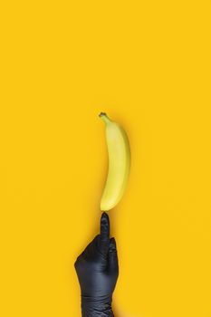 Hand in black glove holds banana on yellow background. Food art. Conception coronavirus new lifestyle concept.
