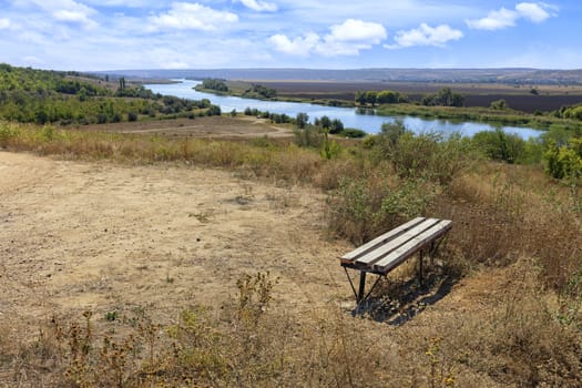 Autumn panorama landscape, sunny day. The edge of a country road near a wooden bench, which stands on a high bank of the river.