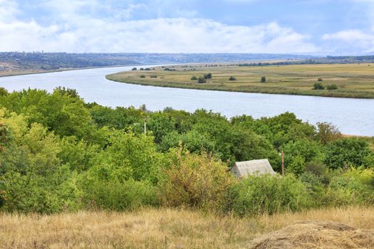 The roof of the farmhouse is immersed in the greenery of the garden against the backdrop of the wide bend of the Southern Bug River and autumn mowed grass in the foreground.