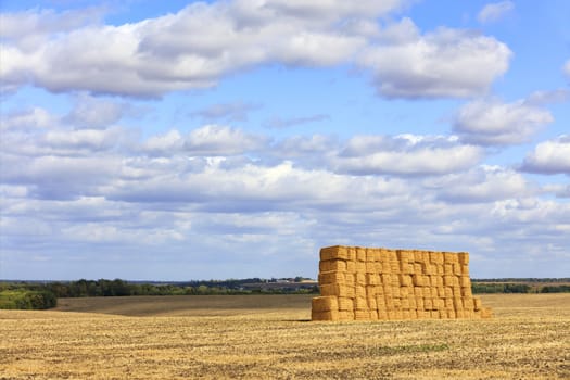 A large straw crop is neatly stacked in the middle of the field after harvesting wheat and glistens in the sun against a blue cloudy sky.