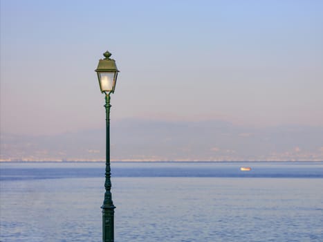 A street lamp is lit by the rays of the setting sun against the backdrop of the sea bay.