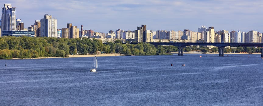 Sailing yacht goes along the banks of the Dnipro River against the backdrop of urban residential areas and a bridge over the river.