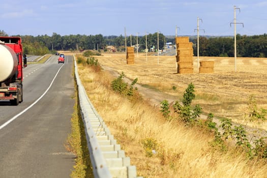 Rural landscape of an asphalt road near a harvested field on an autumn day. Landscape with a field, stacks of straw and a busy road among the hills.