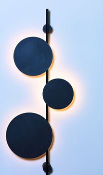 Stylish vertical metal wall lamp in the form of three celestial bodies in an eclipse against a blue wall makes soft warm lighting.