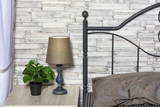 Calm pastel beige with a brown tone of the bed, a green flower pot, an elegant aged table lamp on the bedside table go well with the gray wall of old wooden boards.