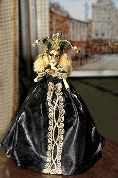 Doll of young Halloween witch in a black evening dress with dark make-up on her face and a spreading hat with a cocked hat on her head on a blurred background of the old city.