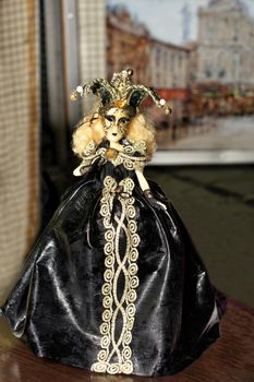 Doll of young Halloween witch in a black evening dress with dark make-up on her face and a spreading hat with a cocked hat on her head on a blurred background of the old city.