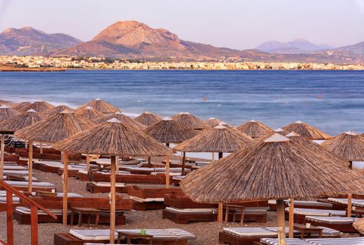 Thatched peaks of beach umbrellas with deck chairs on the deserted beach of the sea promenade against the backdrop of the rays of the setting sun on the horizon with the city of Corinth in Greece, mountain ranges in haze and blur.