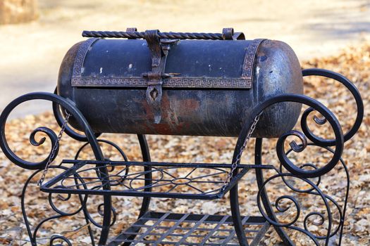 Nice old metal brazier with twisted curly handles made of wrought iron with a beautiful pattern on the background of autumn fallen leaves on a sunny day in blur.