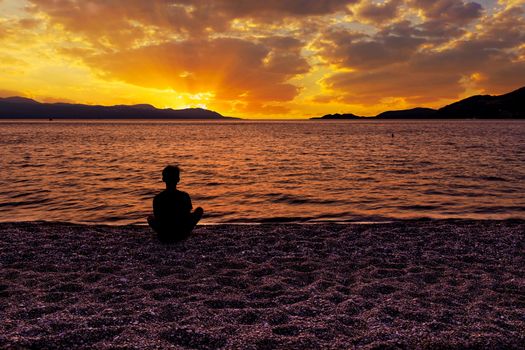 Dark silhouette of a teenager who sits at dusk on a beach of small pebbles on the seashore and watches a beautiful, bright sunset on the Gulf of Corinth, Loutraki, Greece, image with copy space.