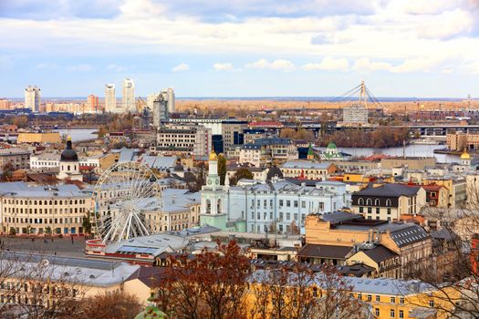 View of the old Podolsky district of the city of Kyiv, the old square with a Ferris wheel and a bell tower with a gilded dome, the Dnipro River and the North Bridge through it.