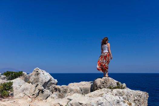Young woman in a colorful skirt and white top stands on the edge of a cliff and looks at a flat blue sea horizon. Seascape. Rock edge. A lot of air, image with copy space.