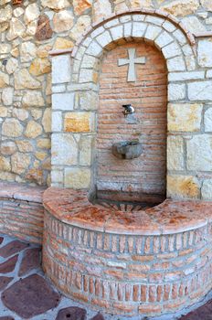 Brass metal faucet sticks out of the stone arch wall of the St. Potapius Convent near Loutraki, spring water flows in a slow stream into a stone bowl.