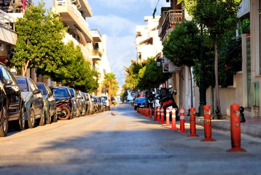 Red reflective road columns separate the pedestrian area of the road from the carriageway amid parked cars, a deserted street and bright morning sunlight in blur.