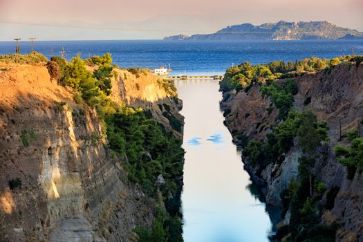 The Corinth Canal in the morning summer day illuminates the bright rising sun of Greece, a view of the Gulf of Corinth from the height of a pedestrian bridge, the channel connects the Saronicos of the Aegean Sea and the Corinthian Gulf of the Ionian Sea.
