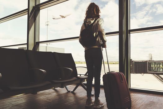Vacation travel concept - rear view of a young girl with a suitcase waiting for the departure of her plane on the background of the airport ( vintage effect)