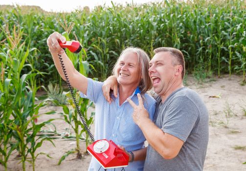 two elderly men take a comic selfie by retro phone in corn field on sunny summer day