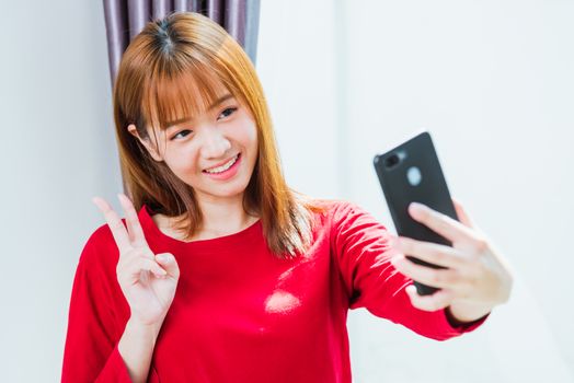 Work from home concept, Asian young woman smiling, student girl sitting on chair make video call or selfie photo from smart mobile phone during working with laptop computer at home office