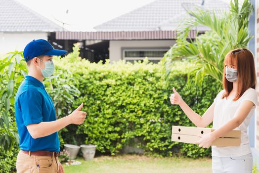 Asian delivery courier young man giving food pizza box to woman customer receive both protective face mask and show thumb up finger for good job, under curfew quarantine pandemic coronavirus COVID-19
