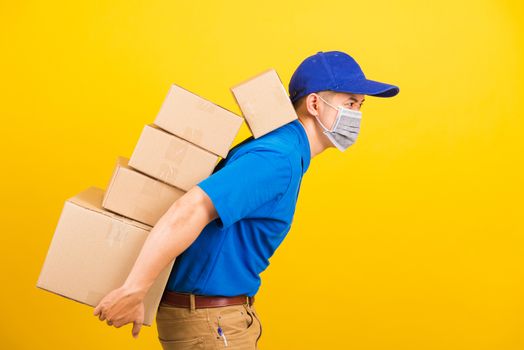 Asian young delivery worker man in uniform wearing face mask protective he has many job lifting stack heavy a lot of boxes on back, under coronavirus COVID-19, studio shot isolated yellow background
