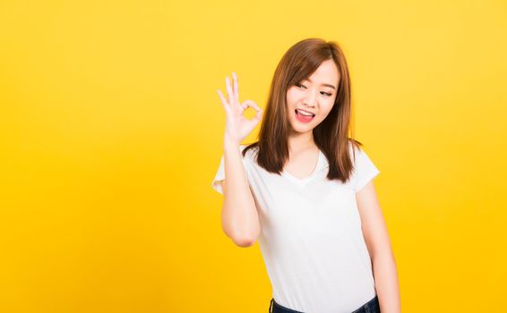 Asian happy portrait beautiful cute young woman teen standing wear t-shirt showing gesturing ok sign with fingers looking to side isolated, studio shot on yellow background with copy space for text