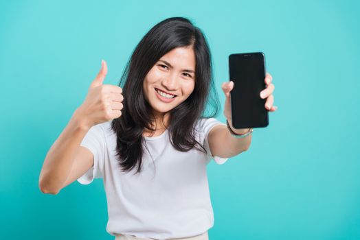 Portrait Asian beautiful happy young woman standing smile, holding blank screen mobile phone and showing thumbs up gesture, shoot photo in studio focus face on blue background, with copy space