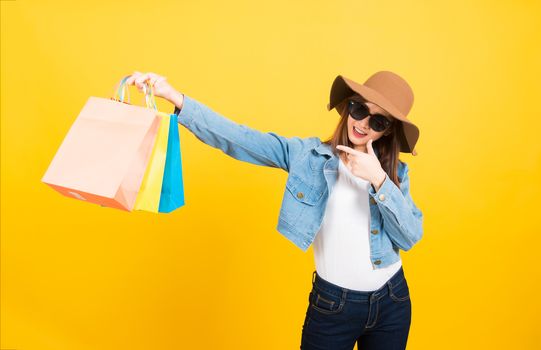 Asian happy portrait beautiful cute young woman smile stand with sunglasses excited holding shopping bags multi color and pointing it looking side isolated, studio shot yellow background by copy space