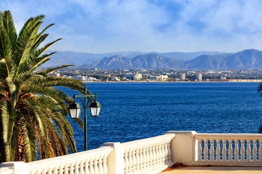 A street lamp under a spreading green palm tree stands near the marble railing and looks at the sea bay, city and mountains in the blur in the rays of bright sunlight, image with copy space.