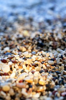Pebble beach on the sea coast is illuminated by a narrow sunbeam and plays with colorful colors, the foreground and background are blurred, close-up, vertical image with copy space.