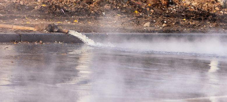 Hot water is pumped out onto the road with a pump and a fire hose, water vapor rises densely above the asphalt, image with copy space.