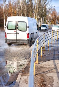 An accident on a heating main, a rupture in a pipe with hot water, trucks driving along a flooded road, water vapor forms a smokescreen over the asphalt, and a metal fence separates the carriageway from the sidewalk.