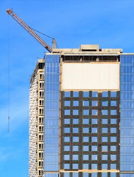 In the construction of modern residential concrete buildings with a glass facade, a tower crane is used, the blue sky is reflected in the windows of the building, vertical image with copy space.