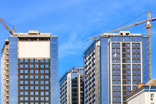 In the construction of modern residential concrete buildings with a glass facade, a tower crane is used, the blue sky is reflected in the windows of the building, horizontal image with copy space.