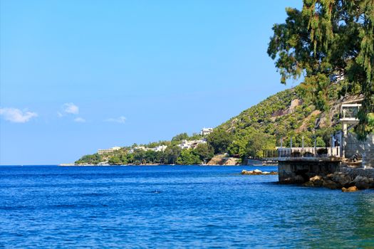 The calm waters of the sea are washed by the rocky green slopes of the coast of the Greek Gulf of Corinth against the background of the blue lagoon, image with copy space.