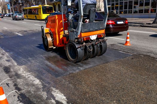 A heavy orange vibratory roller compacts and levels a section of road on a city street fenced with traffic cones, image with copy space.