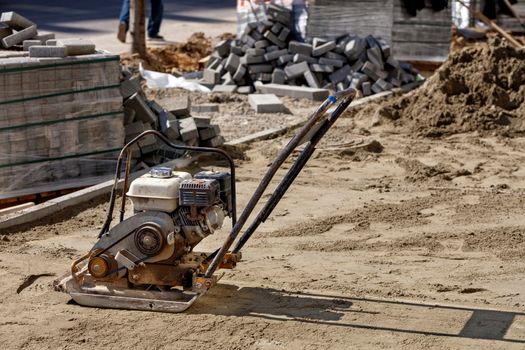 Old worn rusty gasoline compactor stands on the sand against the background of a stack of paving slabs in the blur, image with copy space..