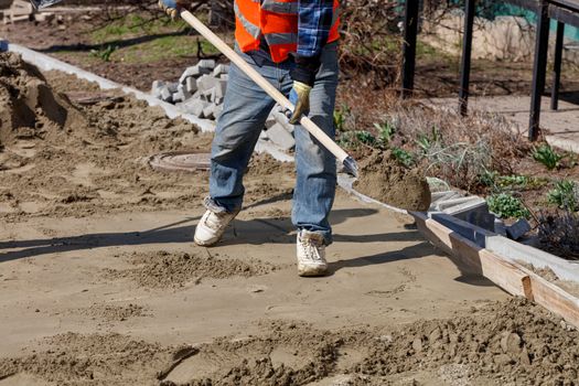 A worker levels the foundation with sand and a shovel under a wooden level for laying paving slabs against the background of the workplace and preparing for work.