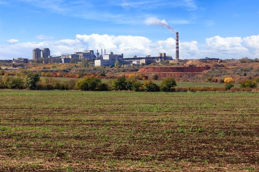 A wide agricultural field after harvesting against the background of a cement plant industrial complex with a quarry on a bright autumn day against the background of a slightly cloudy blue sky.
