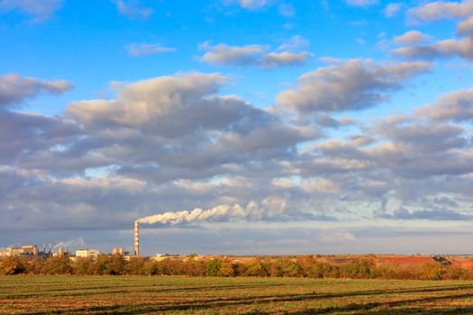 Bright morning autumn sun rays cast long shadows on the agricultural field and illuminate the horizon with the industrial complex and floating clouds in the blue sky.