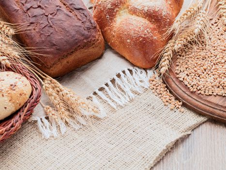 On a wooden table covered with burlap and linen napkins. On a napkin placed rye and wheat bread. Wooden cutting board and basket of bread on a sacking. Ears and grain on a wooden cutting board.