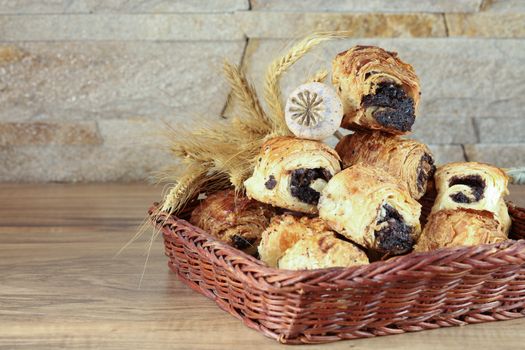Sweet buns with poppy seeds lie in a wicker basket, on a wooden table and near a stone wall - sandstone. Poppy head and spikelets lie on sweet rolls