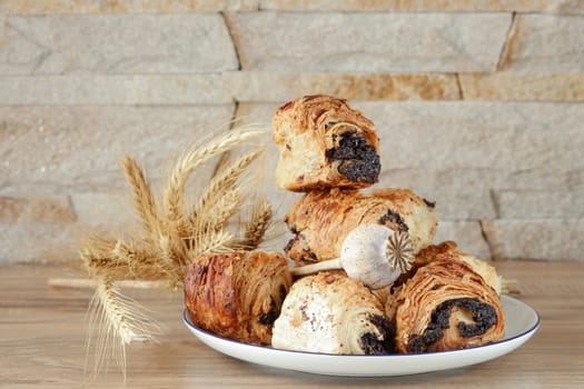 Sweet buns with poppy seeds on a white porcelain plate with a blue rim, on a wooden table and near a stone wall - sandstone. Poppy head and spikelets lie on sweet rolls