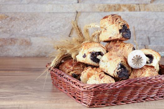 Sweet buns with poppy seeds lie in a wicker basket, on a wooden table and near a stone wall - sandstone. Poppy head and spikelets lie on sweet rolls