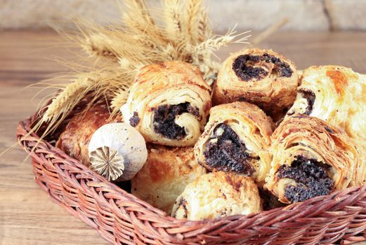 Sweet buns with poppy seeds lie in a wicker basket, on a wooden table and near a stone wall - sandstone. Poppy head and spikelets lie on sweet rolls