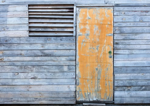Yellow old door and a window with a wooden grate on the facade of a wooden old gray weather-beaten barn.