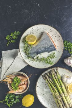 Trout fish  surrounded parsley, lemon, shrimp, prawn, asparagus in ceramic plates. Black concrete table surface. Healthy seafood background.