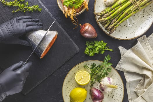 Hands in black gloves cut trout fish on black stone cutting board surrounded herbs, onion, garlic, asparagus, shrimp, prawn in ceramic plate. Black concrete table surface. Healthy seafood background.
