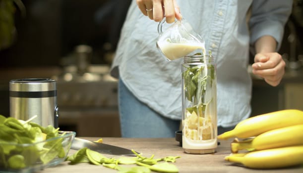 Female hands pouring almond milk in blender glass with banana and spinach leaves. Cooking smoothies in the kitchen. Healthy lifestyle and eating concept